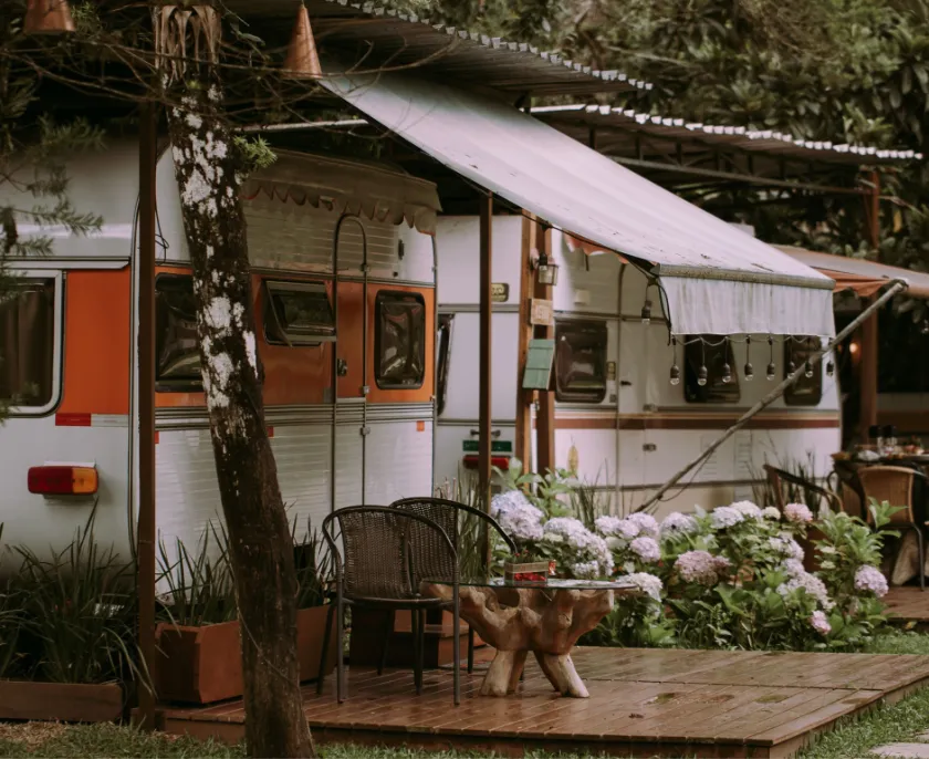 Ein Wohnwagen mit orangefarbenem und weißem Design steht auf einer Holzterrasse, umgeben von grünen Pflanzen und weißen Blüten, mit zwei Stühlen und einem kleinen Tisch darunter einem ausgeklappten Markisentuch.