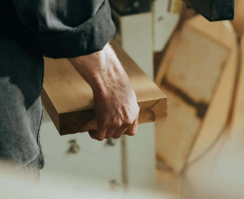Eine Person hält ein Stück Holz in der Hand, im Hintergrund ist Holzwerkstatt mit Werkzeugen unscharf zu sehen.