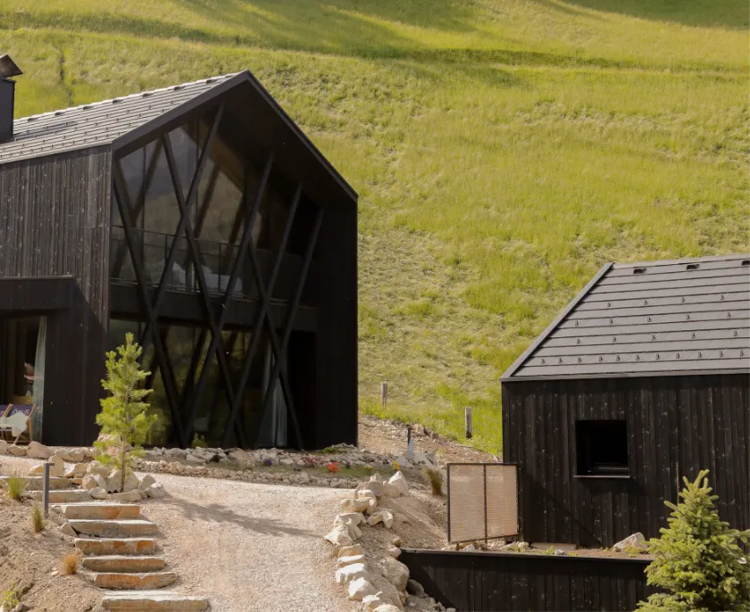 Zwei moderne Hütten mit dunklem Holz an einem Hang mit grünem Gras, mit Steinweg und einigen kleinen Bäumen im Vordergrund.