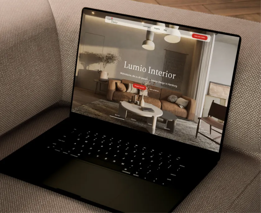 Ein Laptop auf einem grauen Stoffsofa zeigt eine Webseite mit dem Titel "Lumio Interior" und dem Untertitel "Wohnräume, die zu dir passen. Interior Design in Hamburg". Die Webseite zeigt ein modernes Wohnzimmer mit brachem Sofa, rundem Couchtisch, Teppich, Sessel und Deckenlampen.