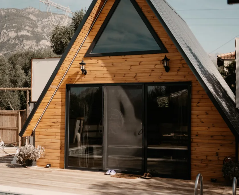 Holzhaus mit A-Rahmen-Design, dreieckigem Oberfenster und großen Glasschiebetüren, davor eine Terrasse mit zwei Schuhpaaren, im Hintergrund Bäume und Berge.