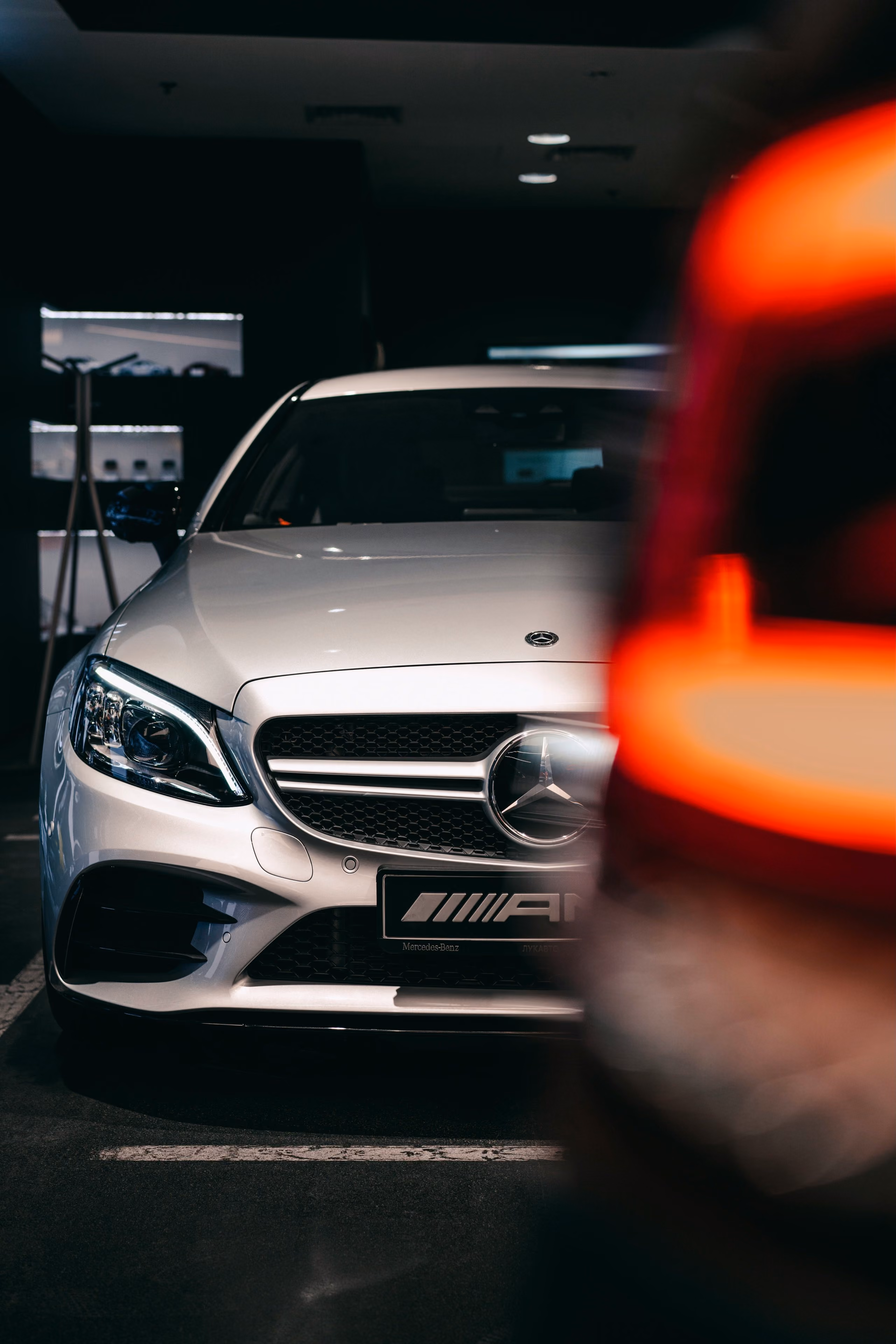Weißer Mercedes-Benz AMG mit beleuchtetem Scheinwerfer und teilverdecktem orangefarbenem Fahrzeug im Vordergrund.