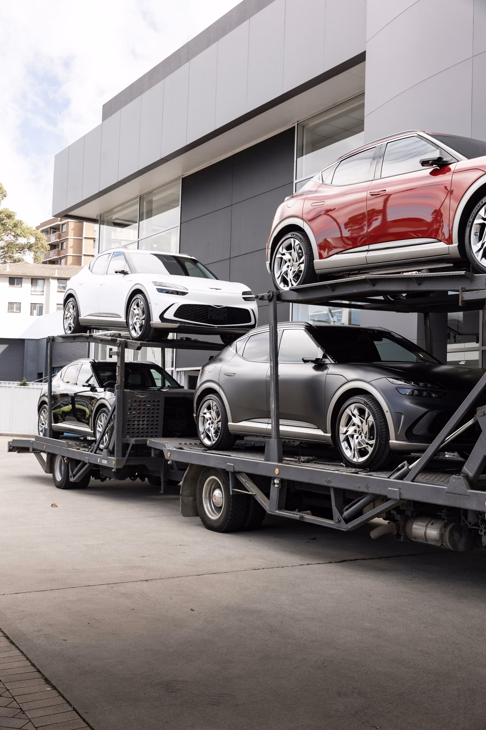 Vier moderne Autos auf einem mehrstöckigen Autotransporter vor einem Gebäude geparkt.
