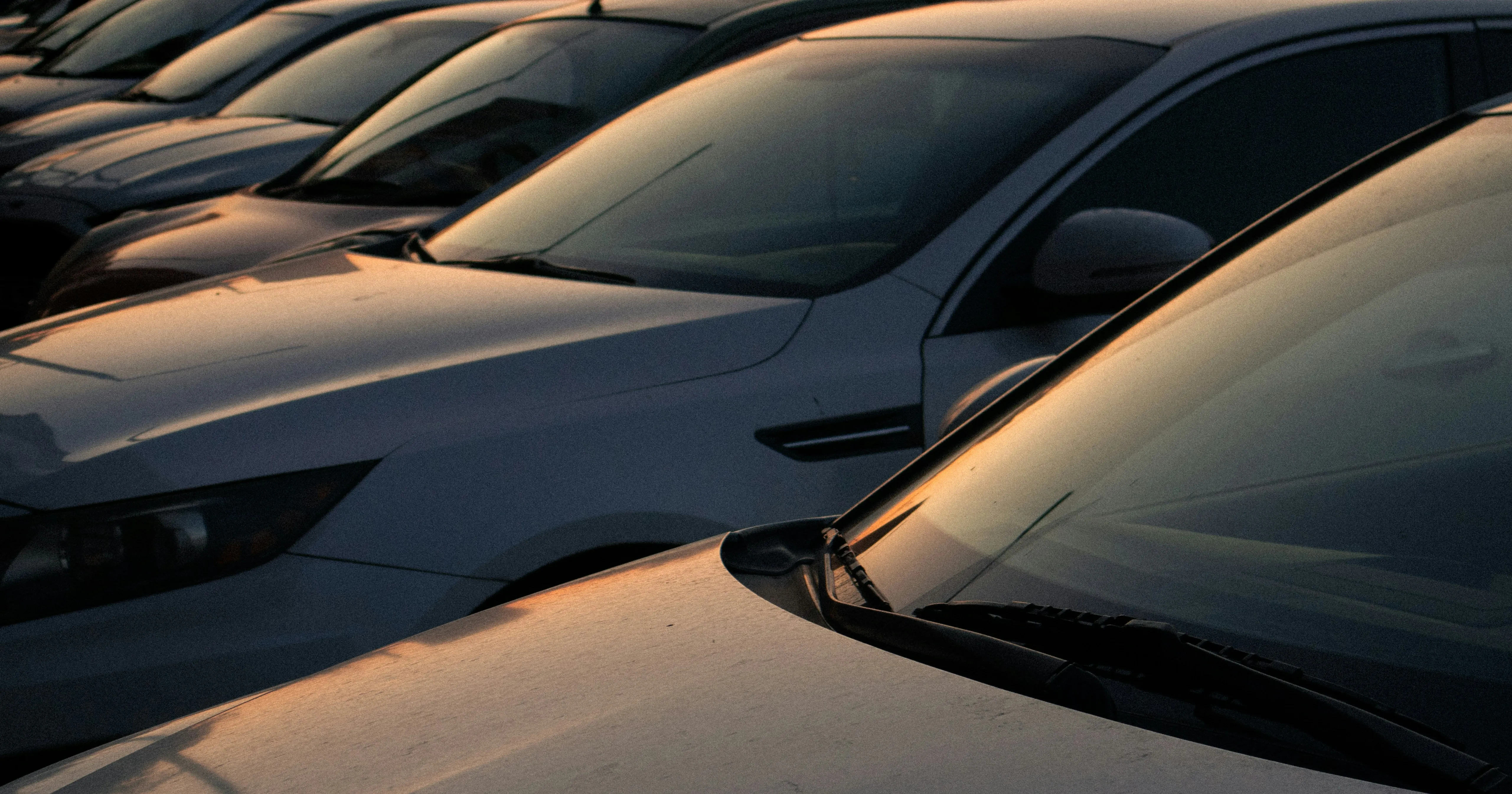 Reihe geparkter Autos bei Sonnenuntergang mit orangefarbenen Reflexionen auf den Windschutzscheiben.