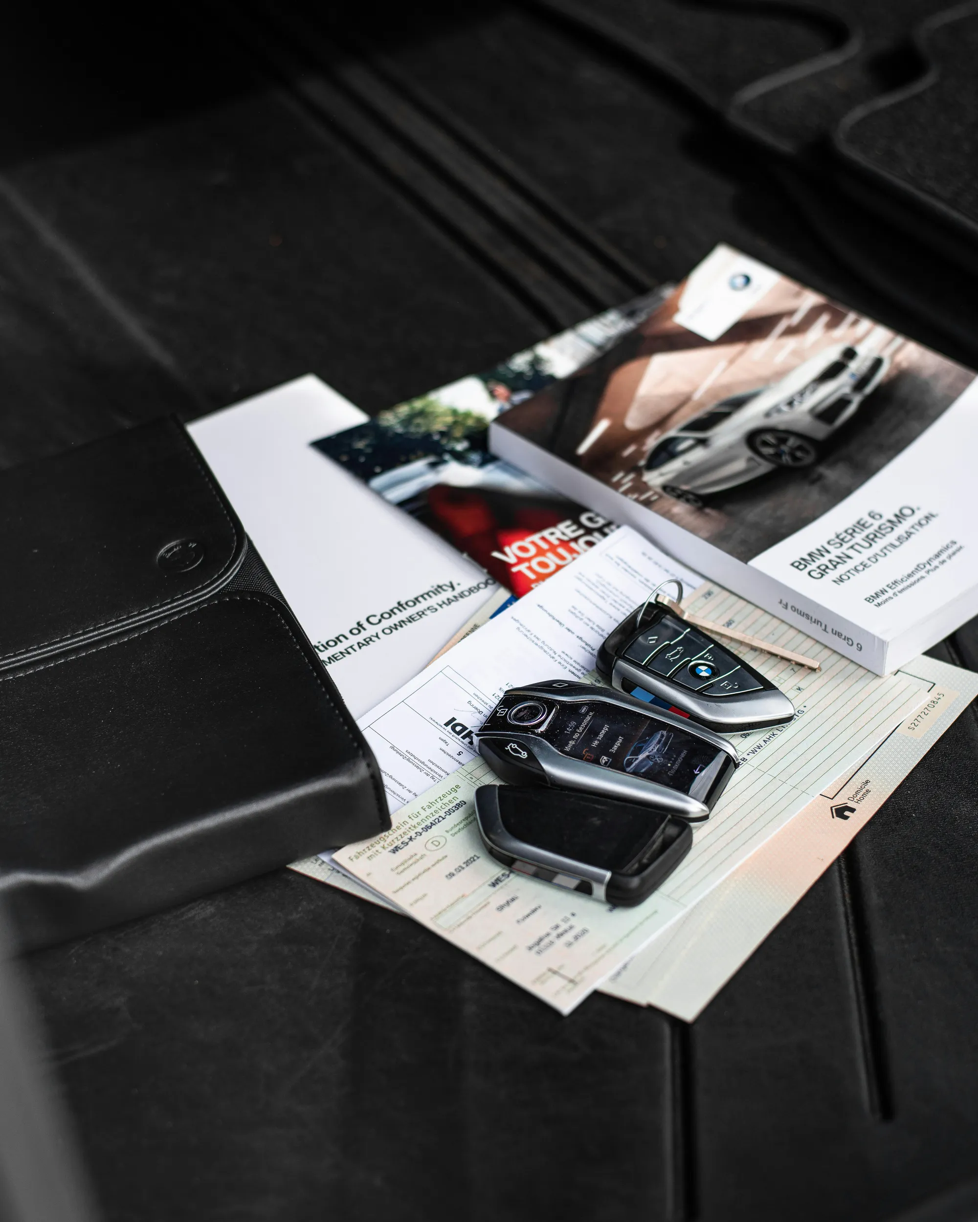 BMW-Autoschlüssel und Fahrzeugpapiere liegen auf einer schwarzen Auto-Matte mit Handbüchern.