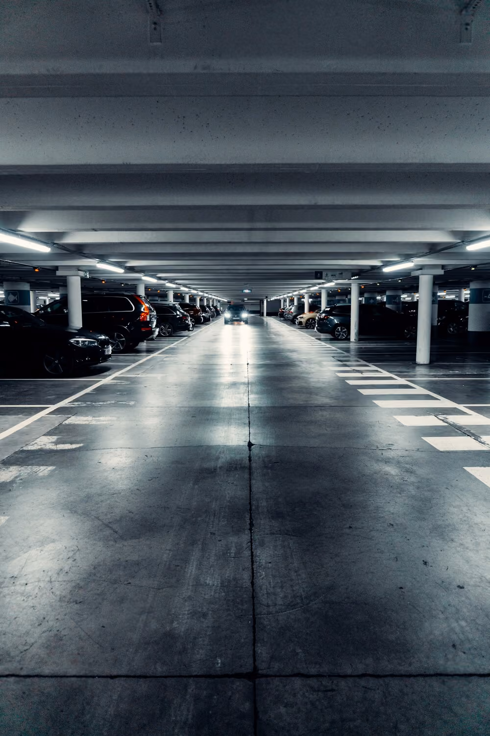 Ein Auto mit eingeschaltetem Licht am Ende eines beleuchteten Parkdecks mit geparkten Autos an beiden Seiten.