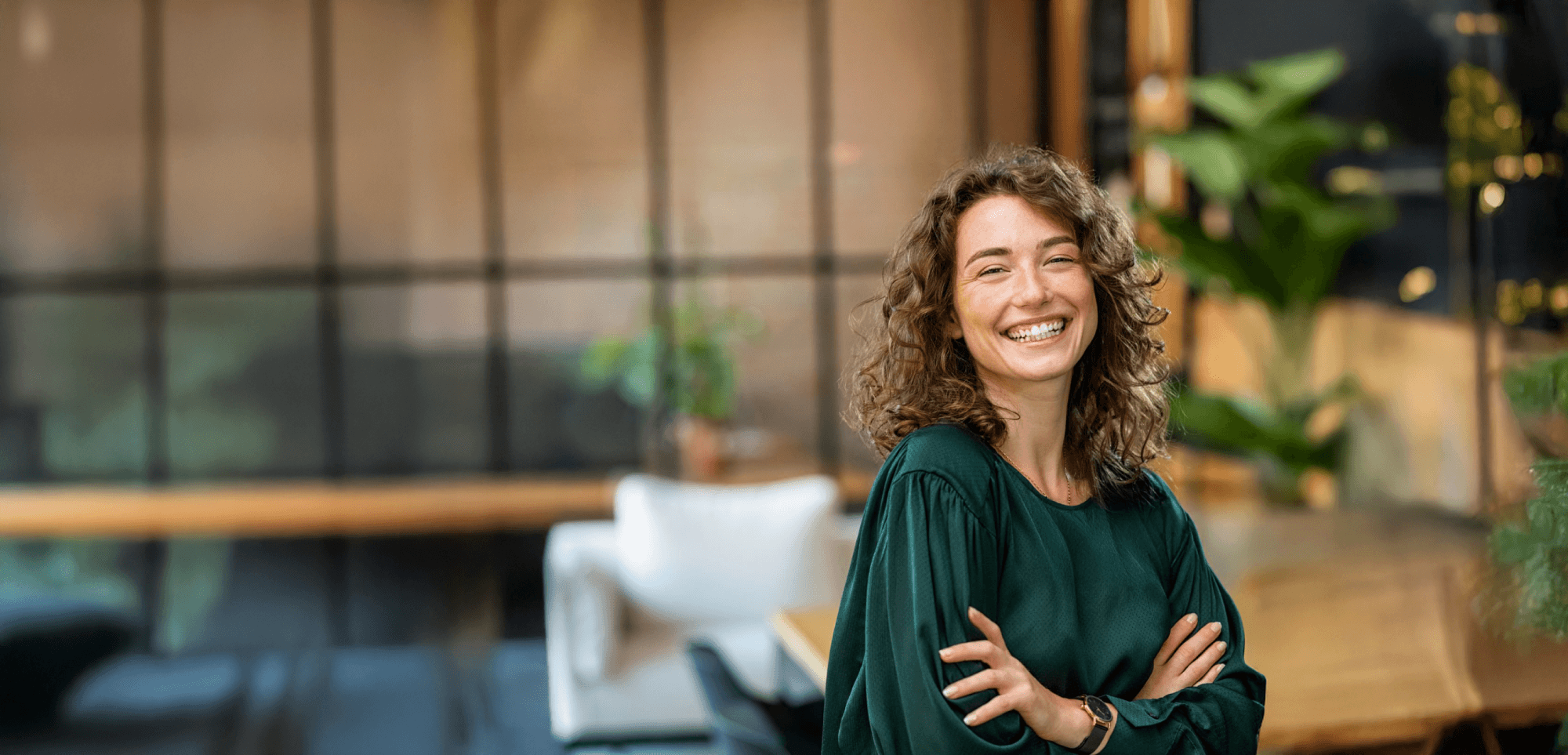 Smiling woman with curly hair and green blouse, arms crossed, in a modern office with plants.