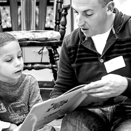Adult reading a book to a young boy while sitting on the floor in a room with wooden chairs.