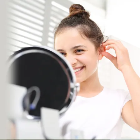 Smiling young woman with hair in a bun looks into a mirror adjusting hair by her ear.