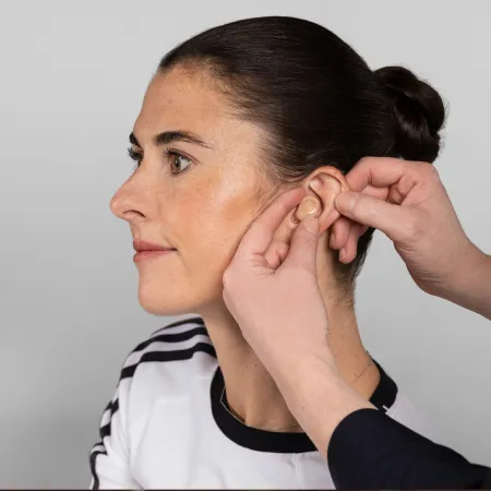 Person in profile has a beige hearing aid placed in their ear by another person's hands.