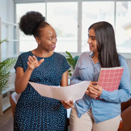 Two women discussing a document in a bright room with large windows and plants.
