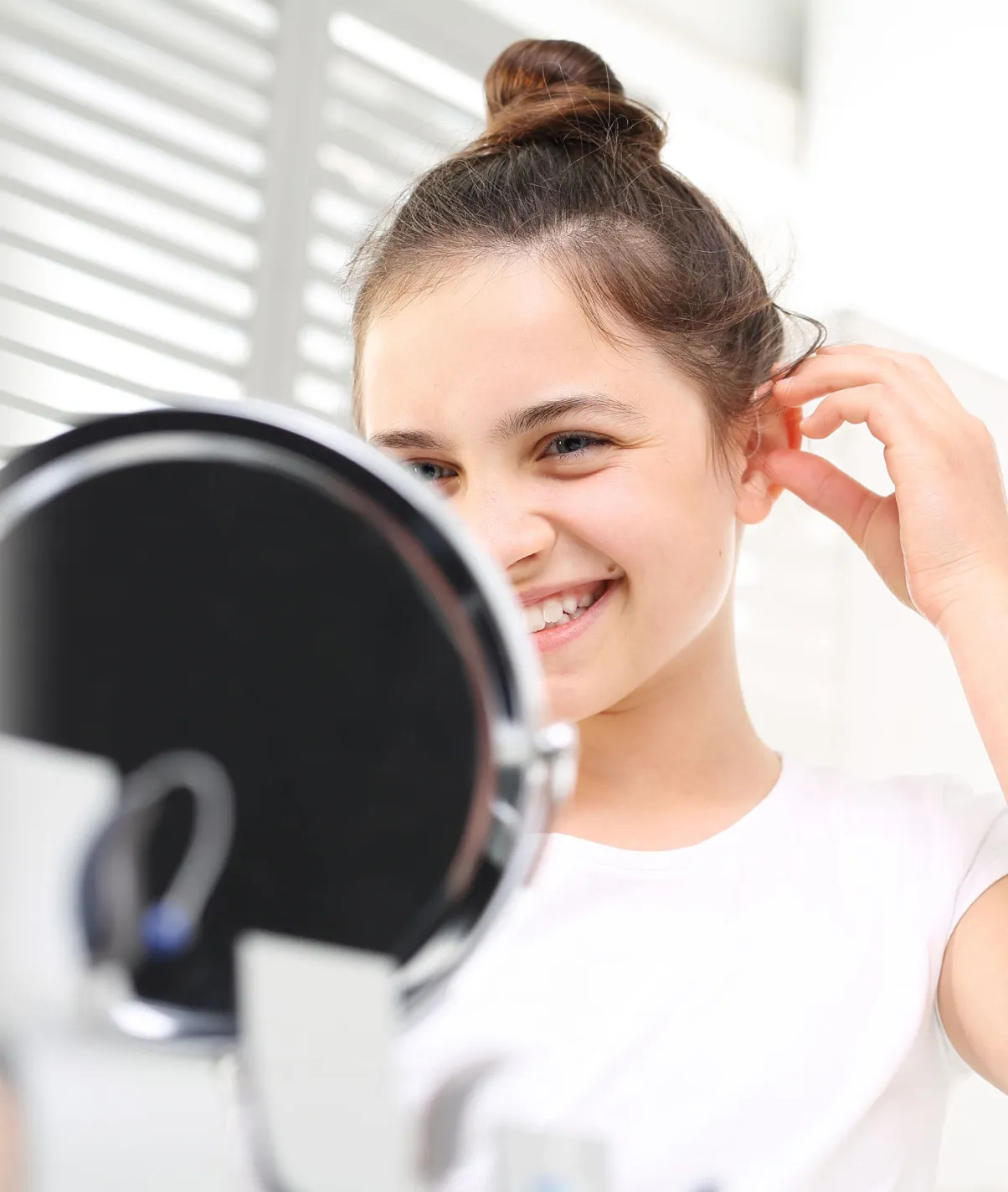 Young woman with hair in a bun smiling and adjusting her ear while looking into a mirror.