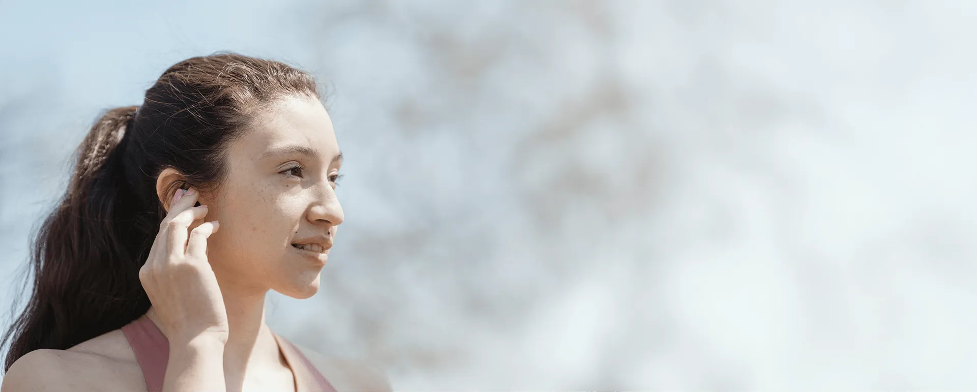 Young woman outdoors adjusting a wireless earbud in her ear with a blurred natural background.