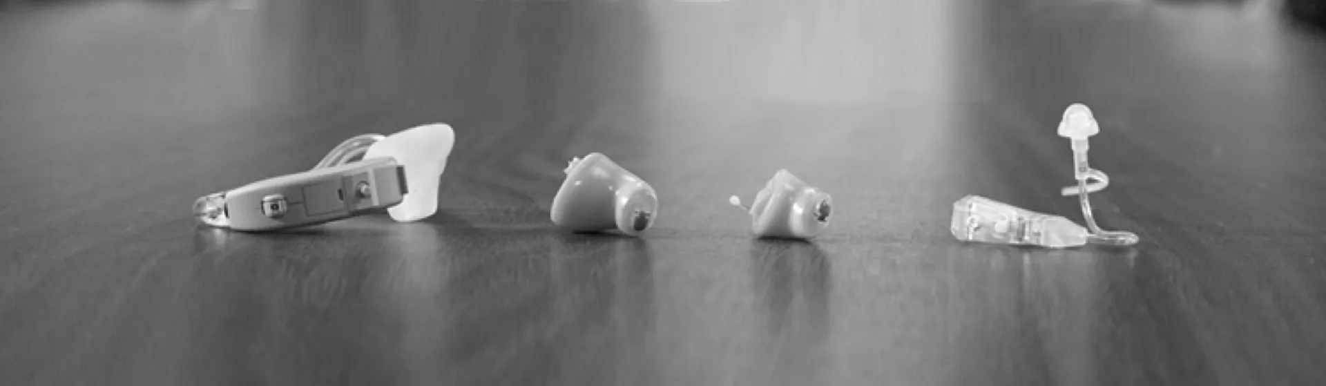 Four hearing aid devices of different styles placed in a row on a wooden surface.