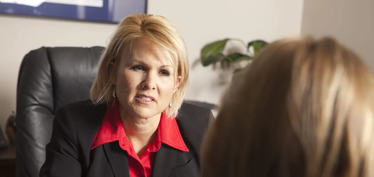 Blonde woman in a red shirt and black blazer sitting in an office chair talking to another person.