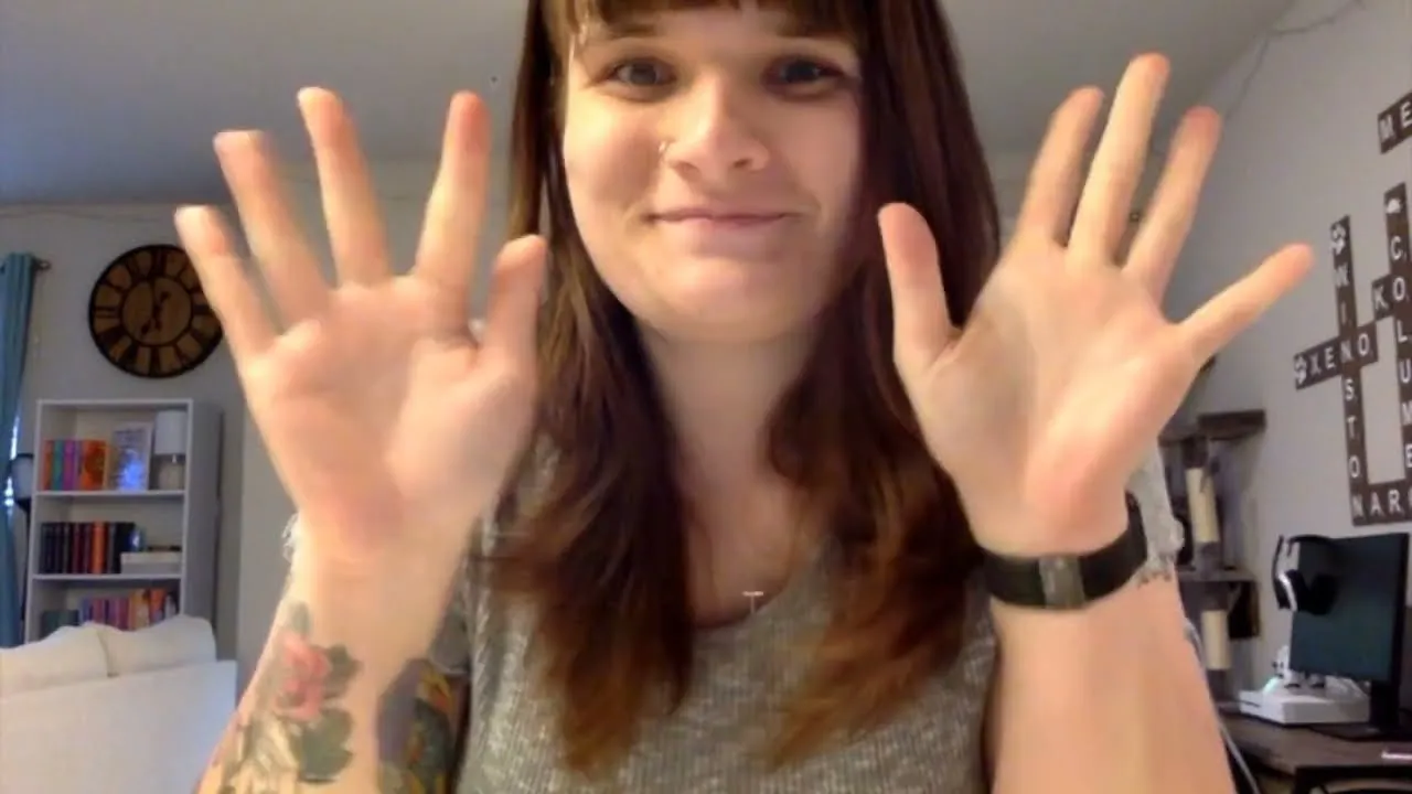 Smiling woman waving both hands with tattoos on arms in a cozy room with books and decor.