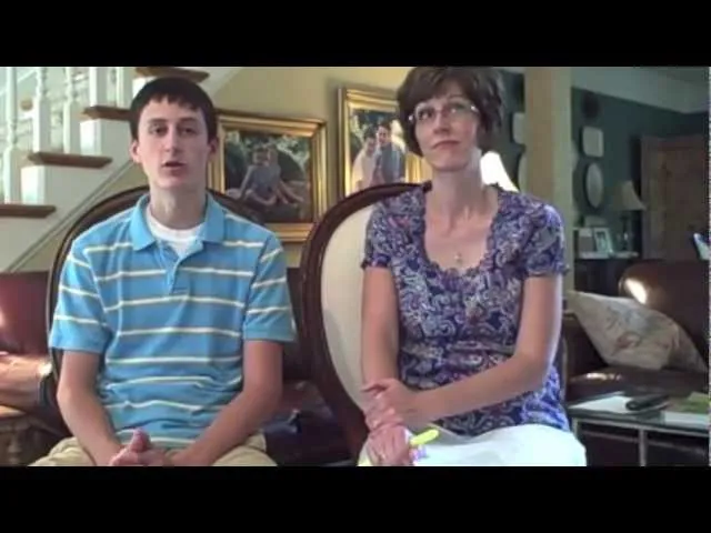 A young man in a striped blue shirt and a woman in a patterned blouse sitting indoors on chairs.