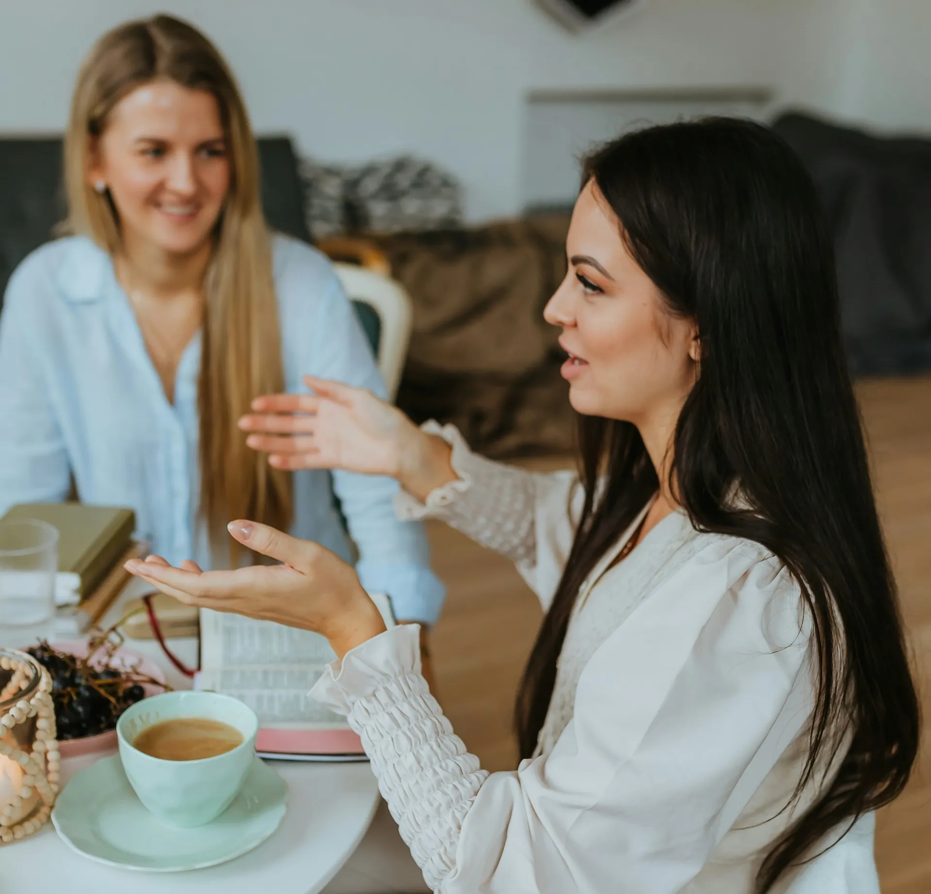 Two women sitting at a table, one talking animatedly with hands raised, the other listening and smiling.