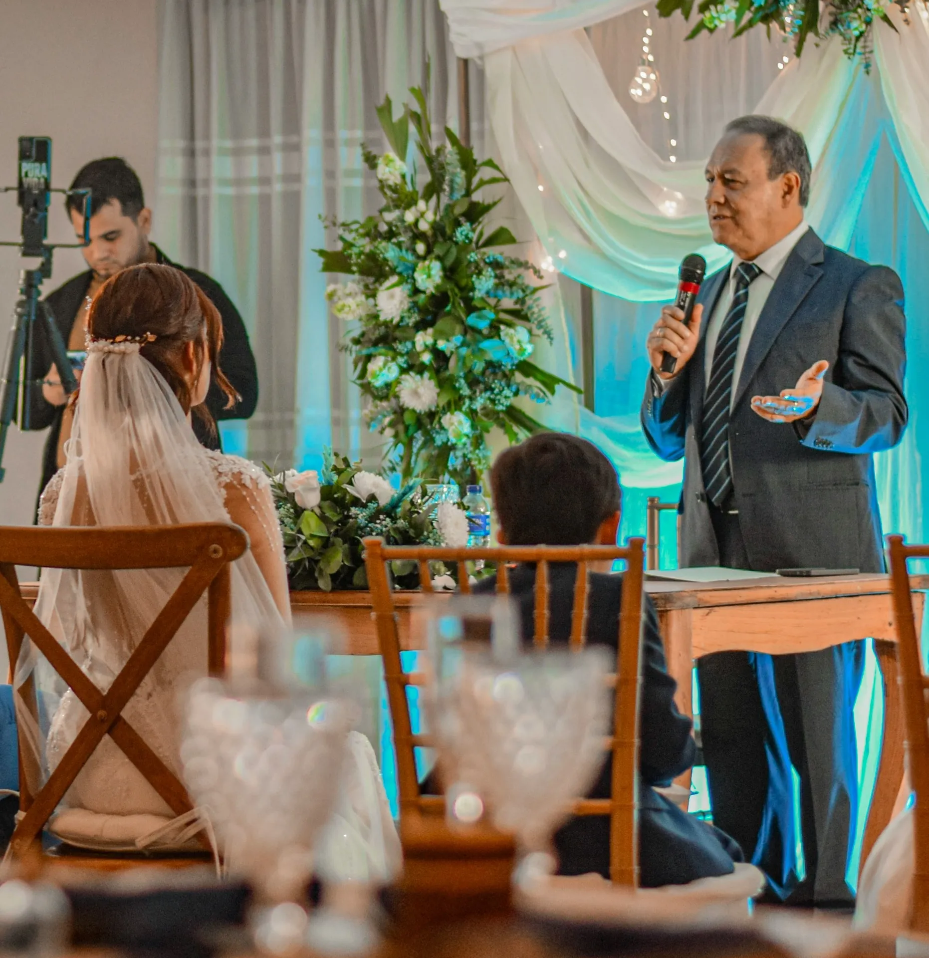 Man in suit speaking into microphone at a wedding, with bride and child seated and floral decoration.