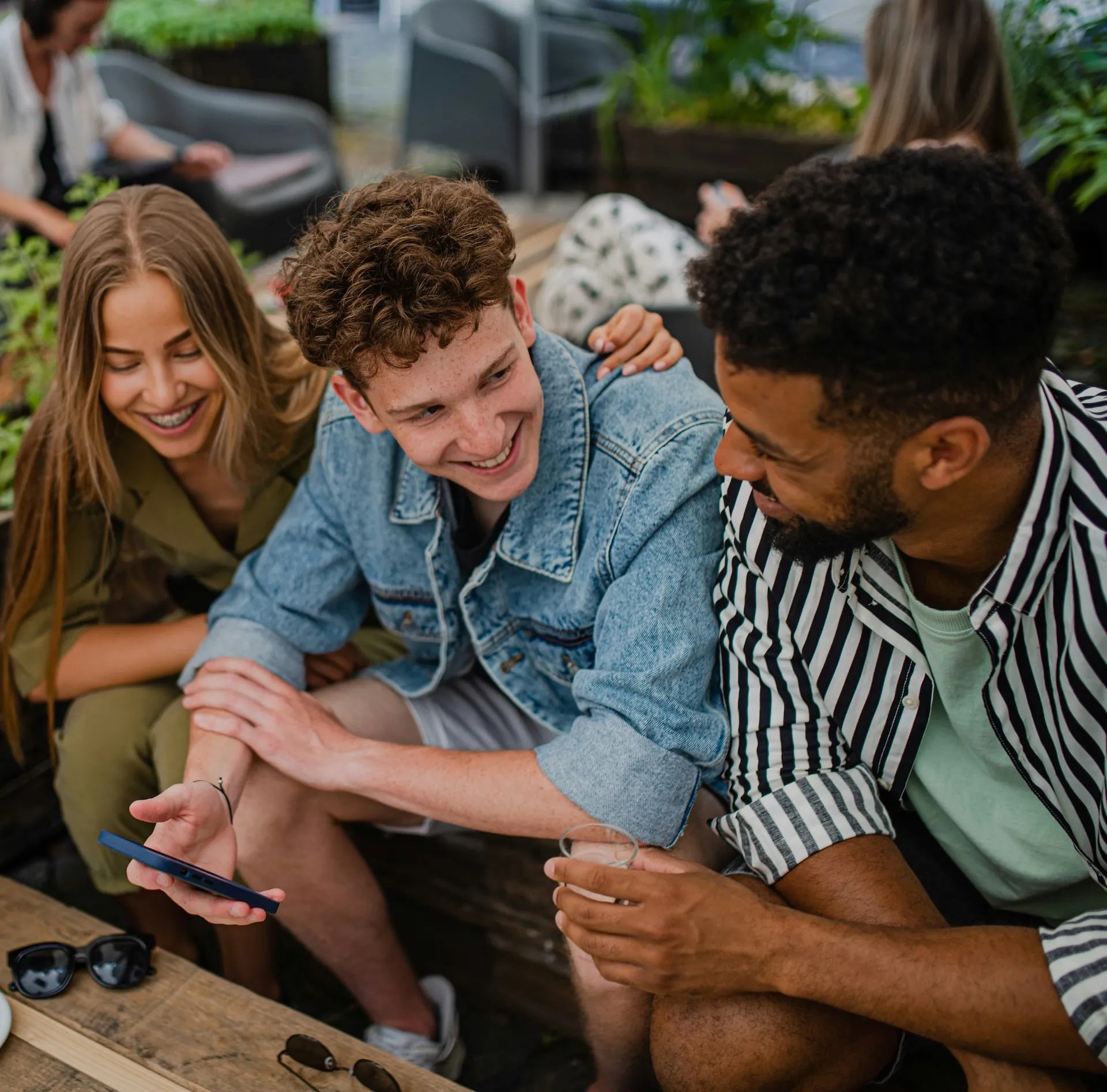 Three friends sitting and smiling, one showing something on a phone while holding a drink.