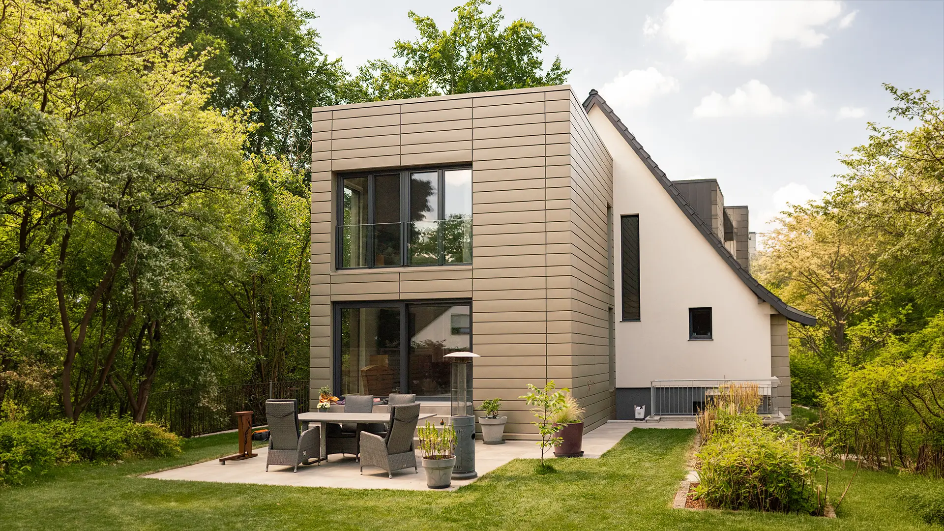 Modern house with large windows, patio set, garden plants, and surrounded by green trees.