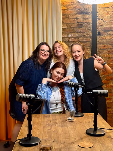 Four women smiling around a wooden table with microphones and drinks in a cozy room with curtain and brick wall.