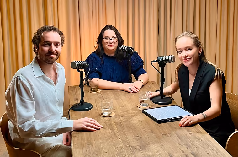 Three people sitting at a wooden table with microphones, recording a podcast or discussion.