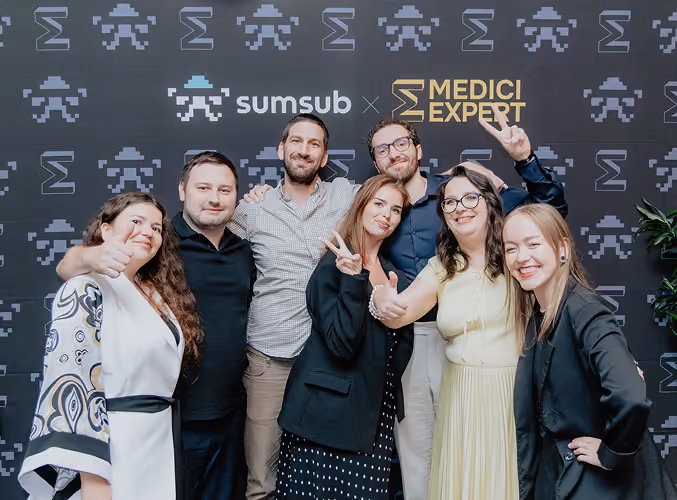 Seven people smiling and posing with peace signs in front of a backdrop with Sumsum and Medici Expert logos.