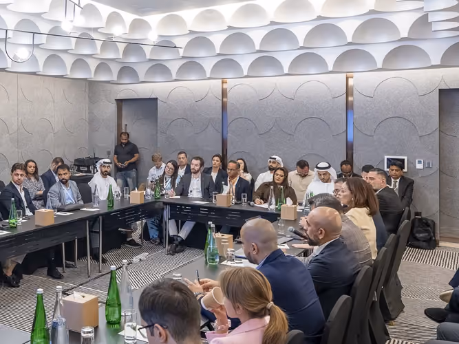 Group of people seated around U-shaped conference tables in a meeting room with modern ceiling design.