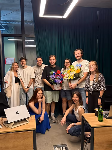 Group of eight smiling people posing indoors, two hold colorful flower bouquets, office setting.