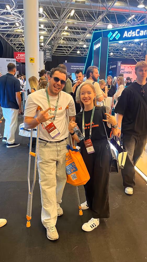 Two smiling people at event, one on crutches and both wearing event badges and casual clothes.