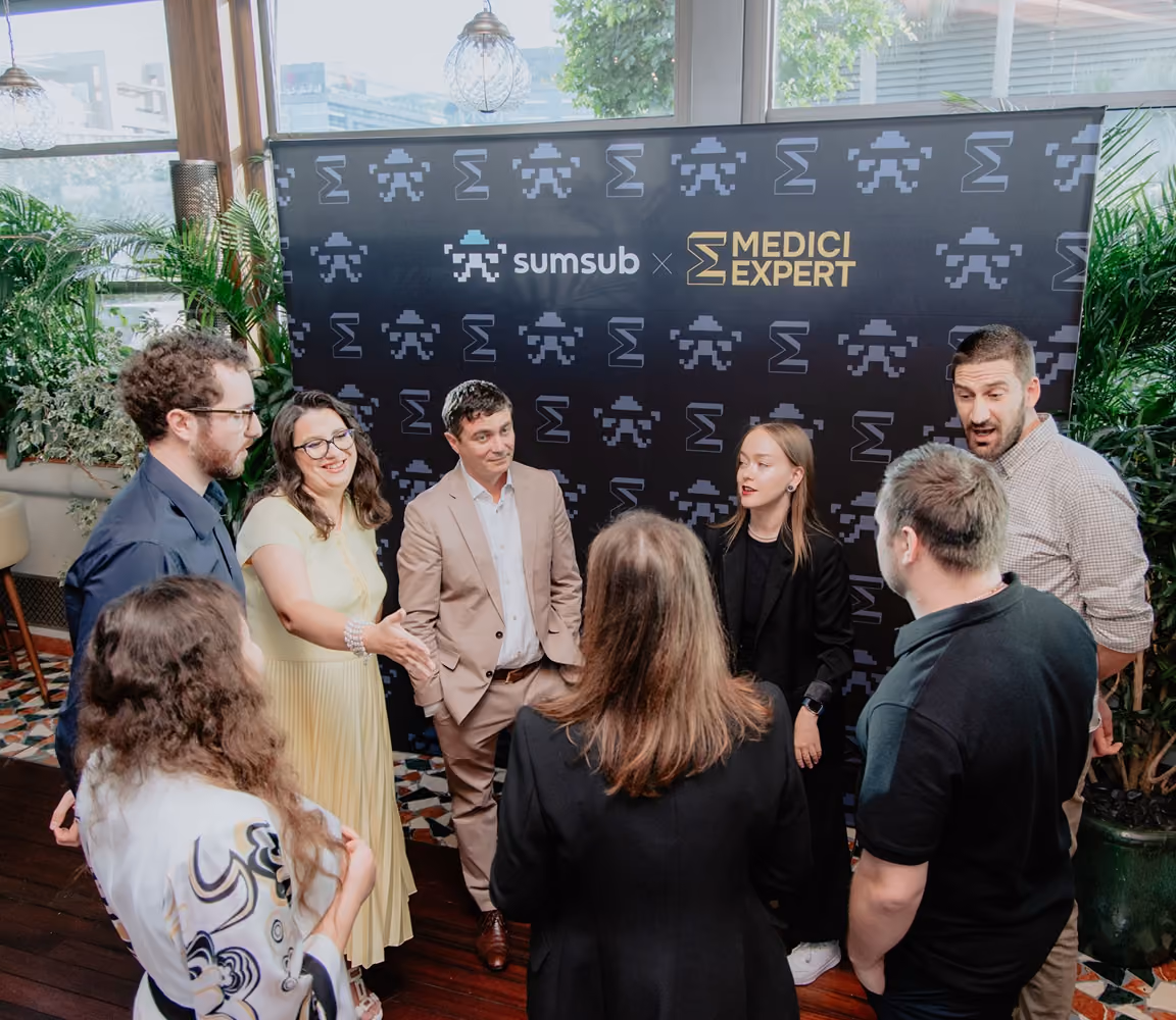 Group of seven people chatting at an event with a sumsub x Medici Expert backdrop.