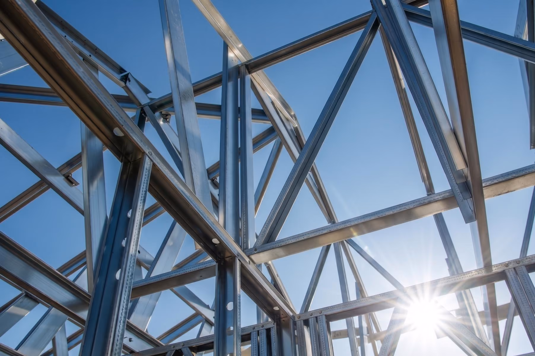 Sunlight shining through steel beams forming a geometric framework against a clear blue sky.