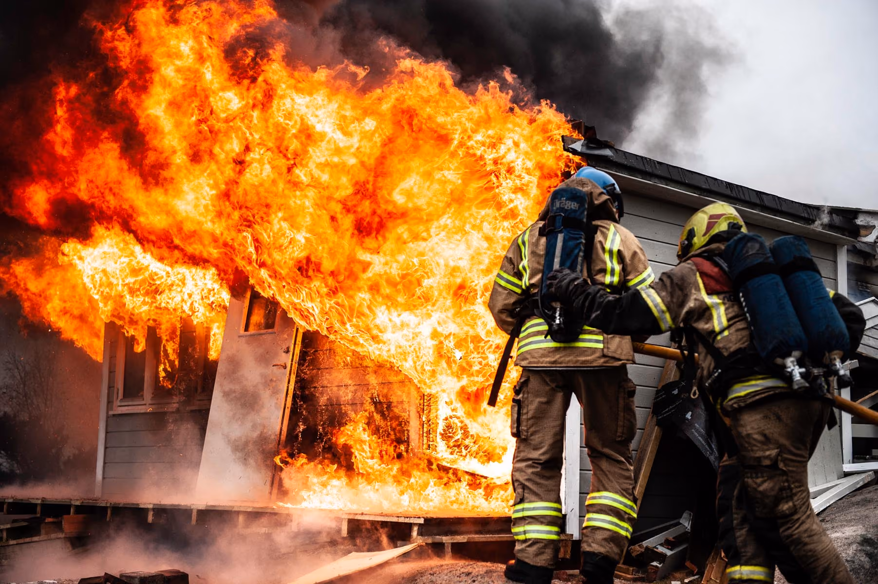 Two firefighters with oxygen tanks approach a house engulfed in large flames and thick black smoke.