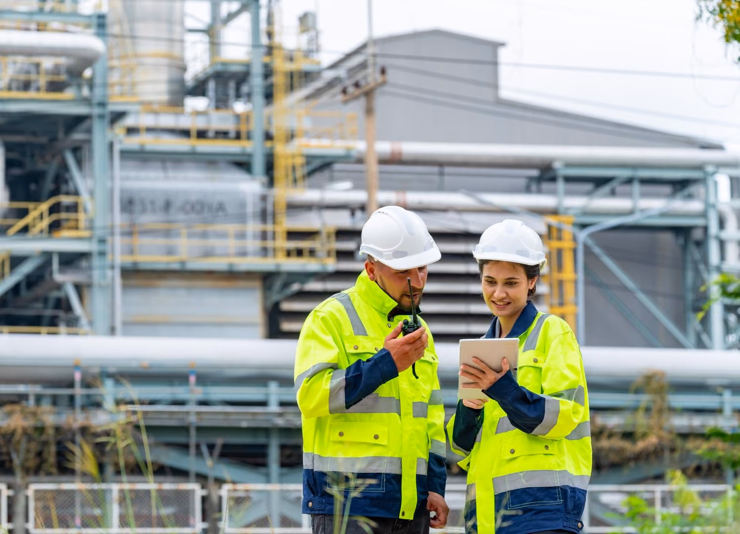 Two construction workers in high-visibility jackets and helmets reviewing a tablet at an industrial site.