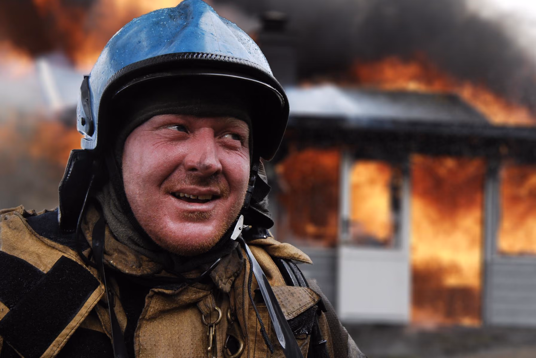 Firefighter in helmet and gear with a burning house engulfed in flames in the background.