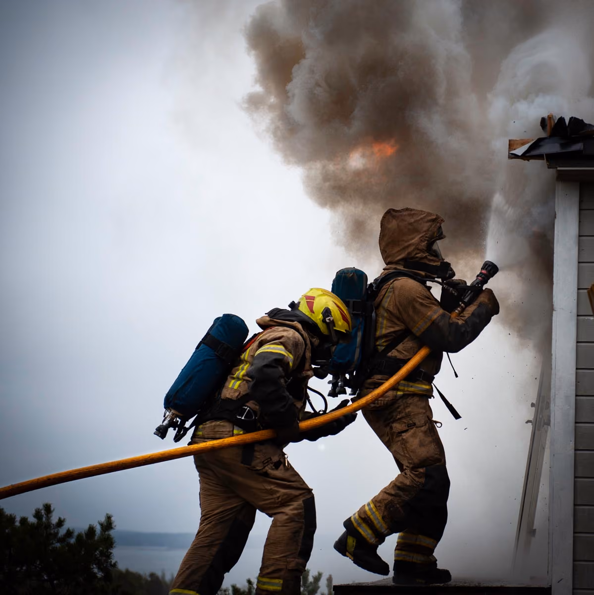 Two firefighters in full gear spraying water on a house fire with thick smoke and flames.