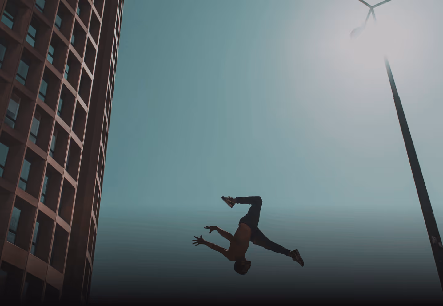Person mid-air performing a backflip outdoors near a building and a street lamp under clear sky.