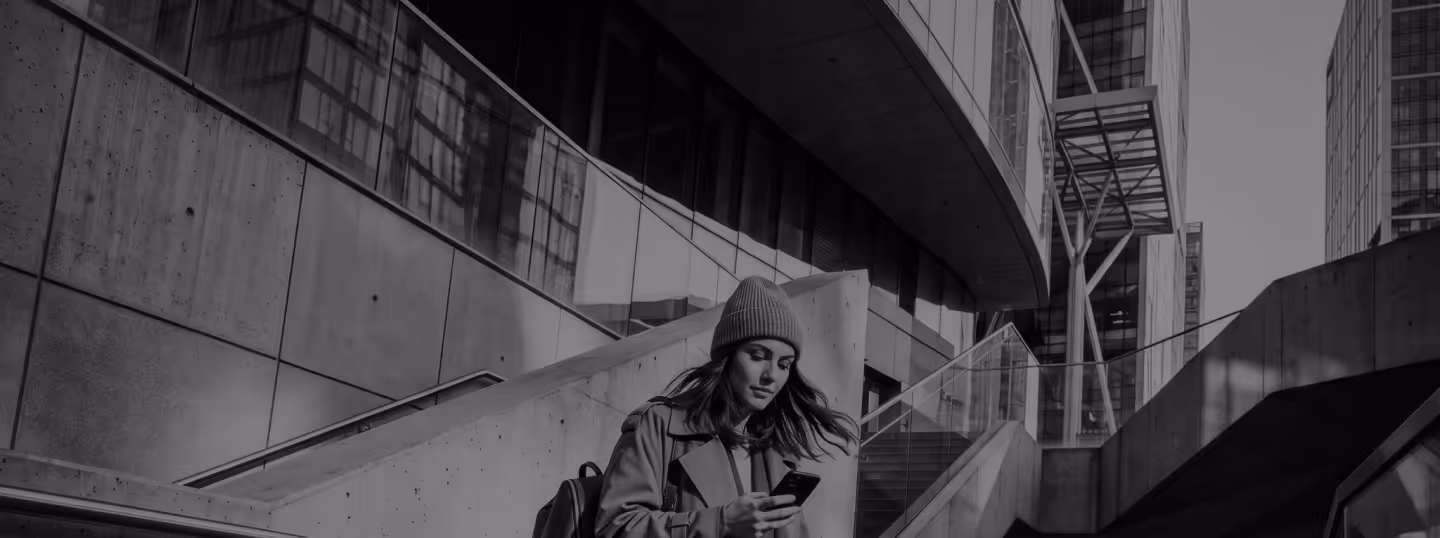 Young woman in beanie and coat looking at phone, standing on urban concrete stairs near modern buildings.