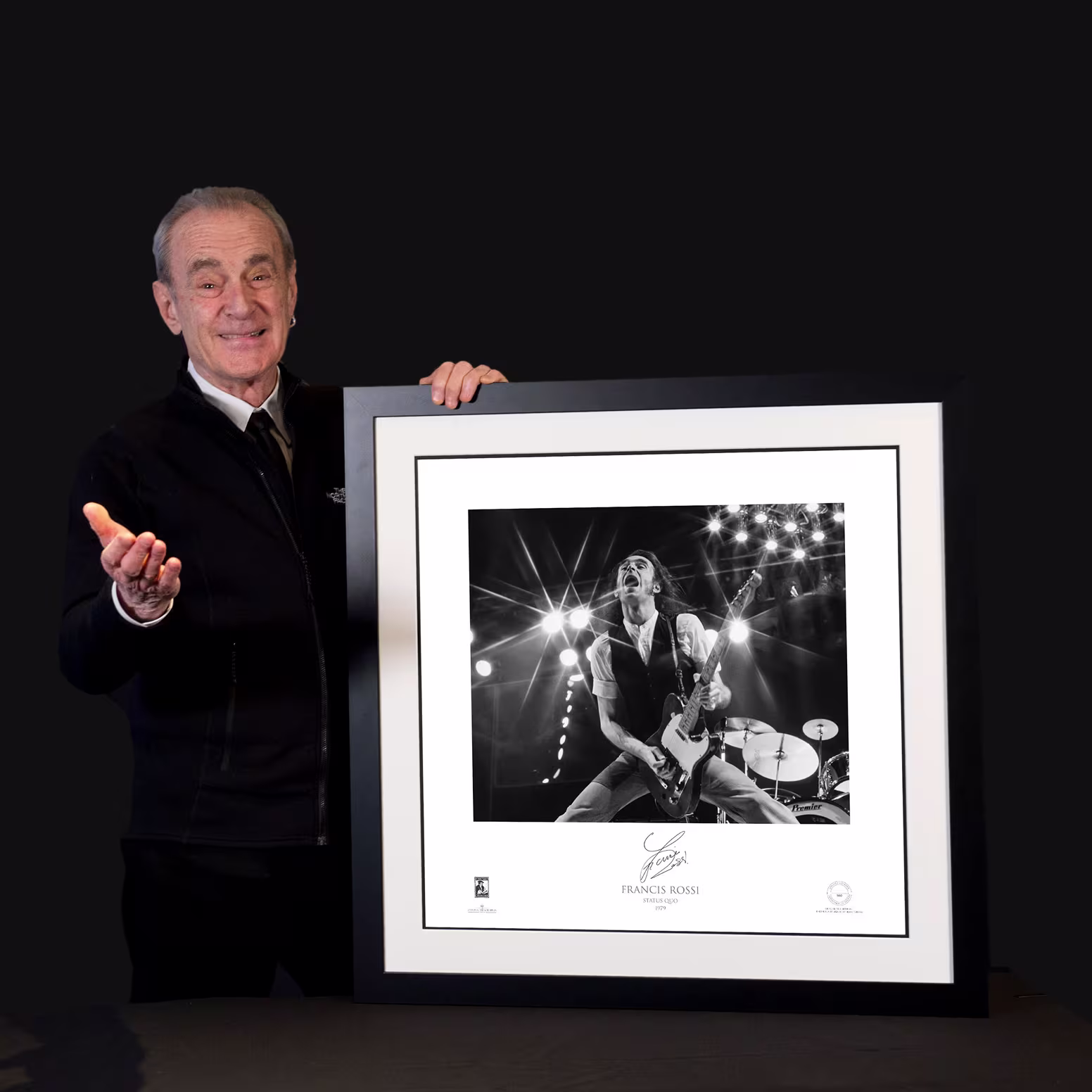 Elderly man presenting a framed black-and-white concert photograph against a black background.