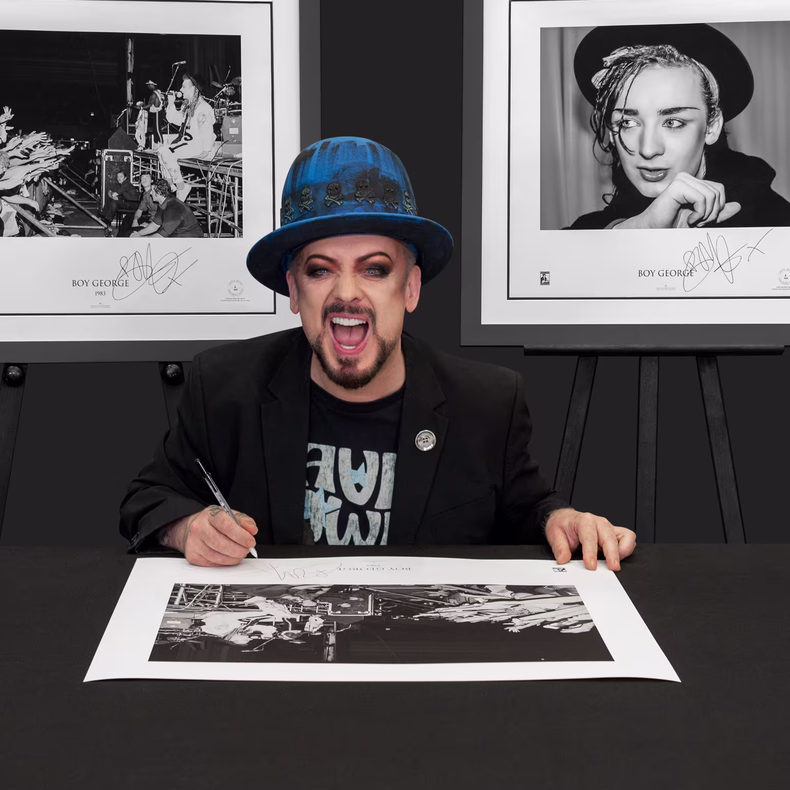 Music artist signing a black-and-white photo print at a table, with framed portraits displayed behind him.