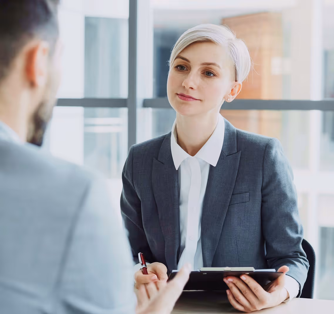 Woman in gray suit listens attentively to a man during a business meeting in a bright office.