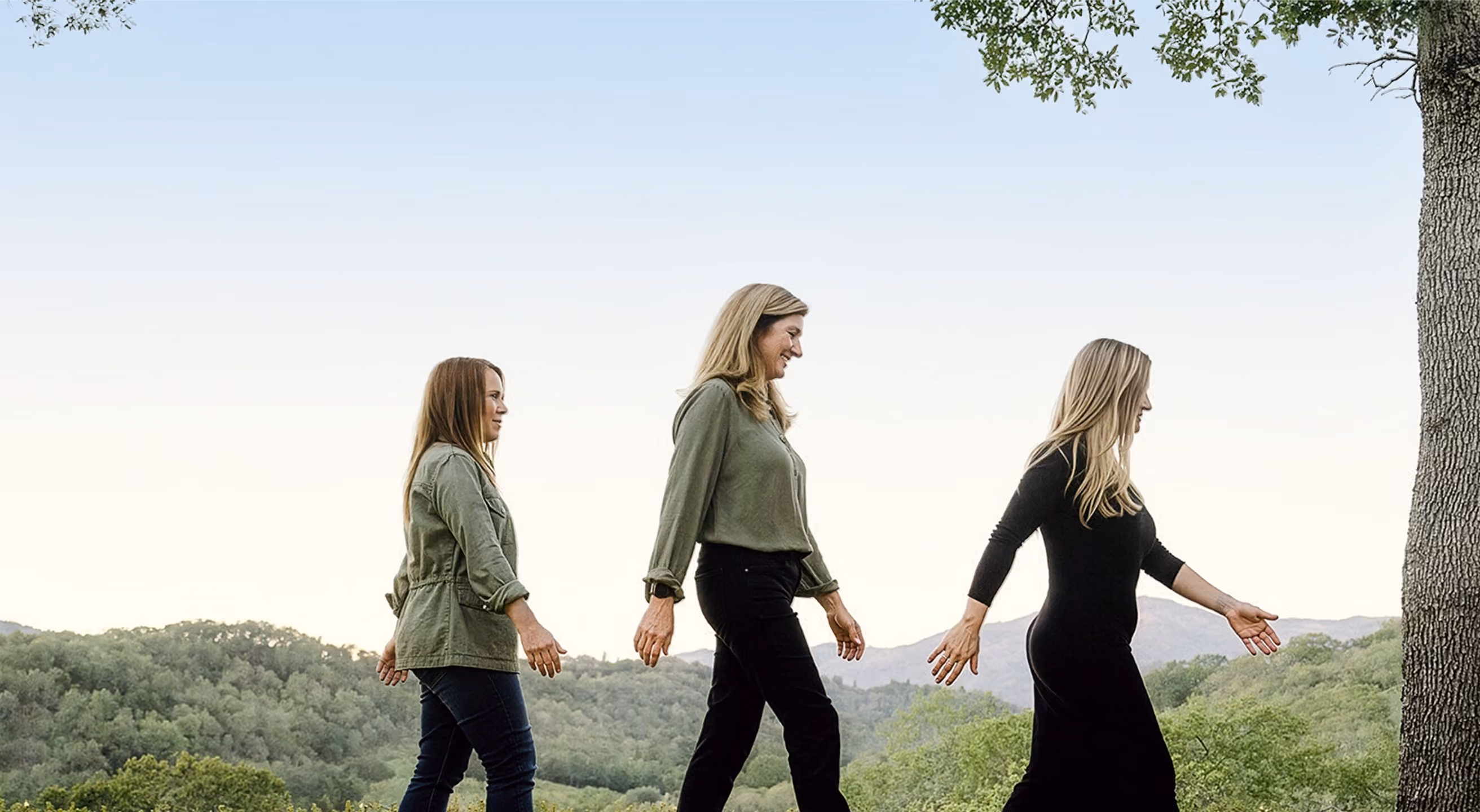 Three women walking in a line outdoors on a curved stone ledge with trees and hills in the background.