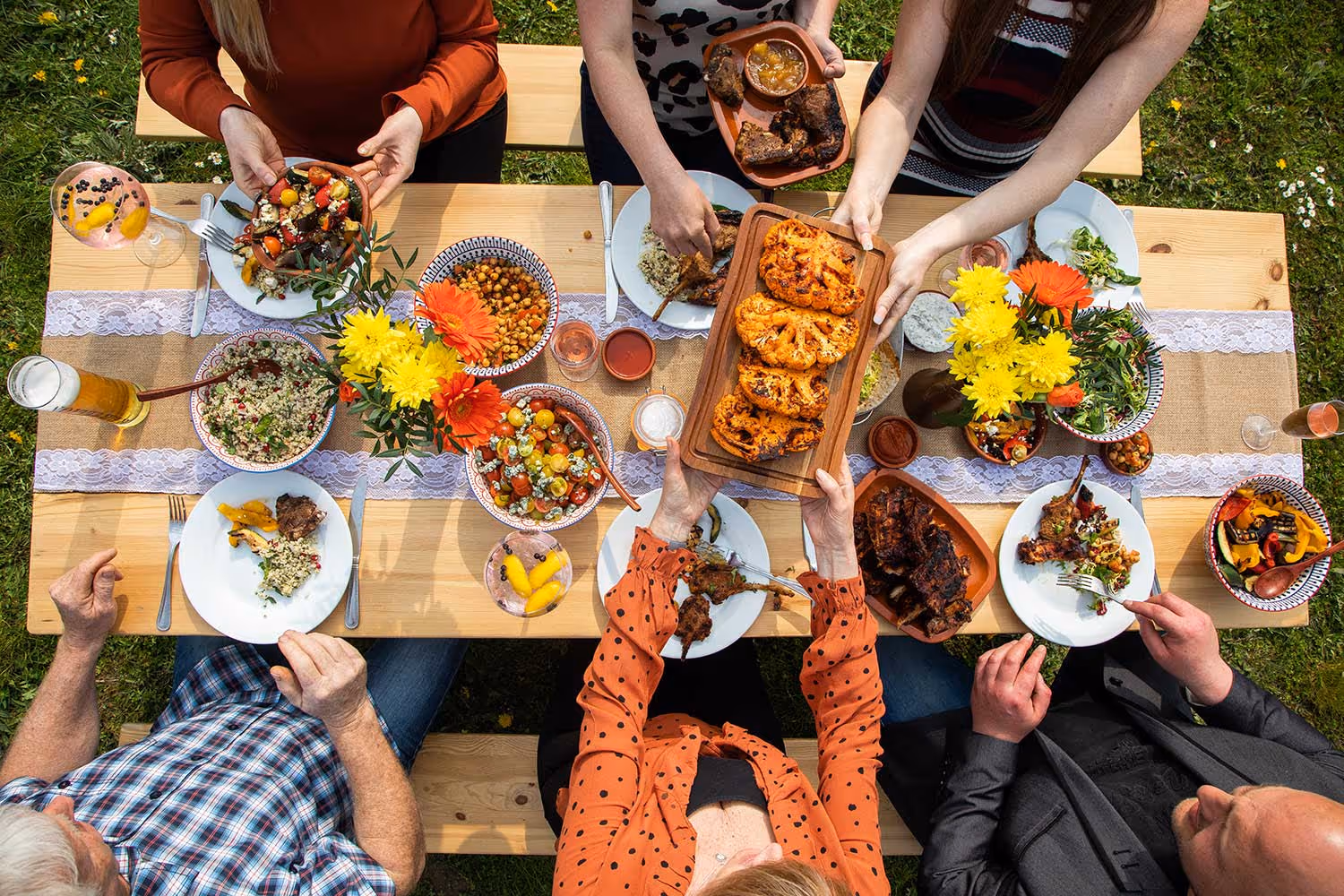 Overhead view of diverse people sharing a colorful outdoor meal with grilled and fresh dishes on a wooden table.