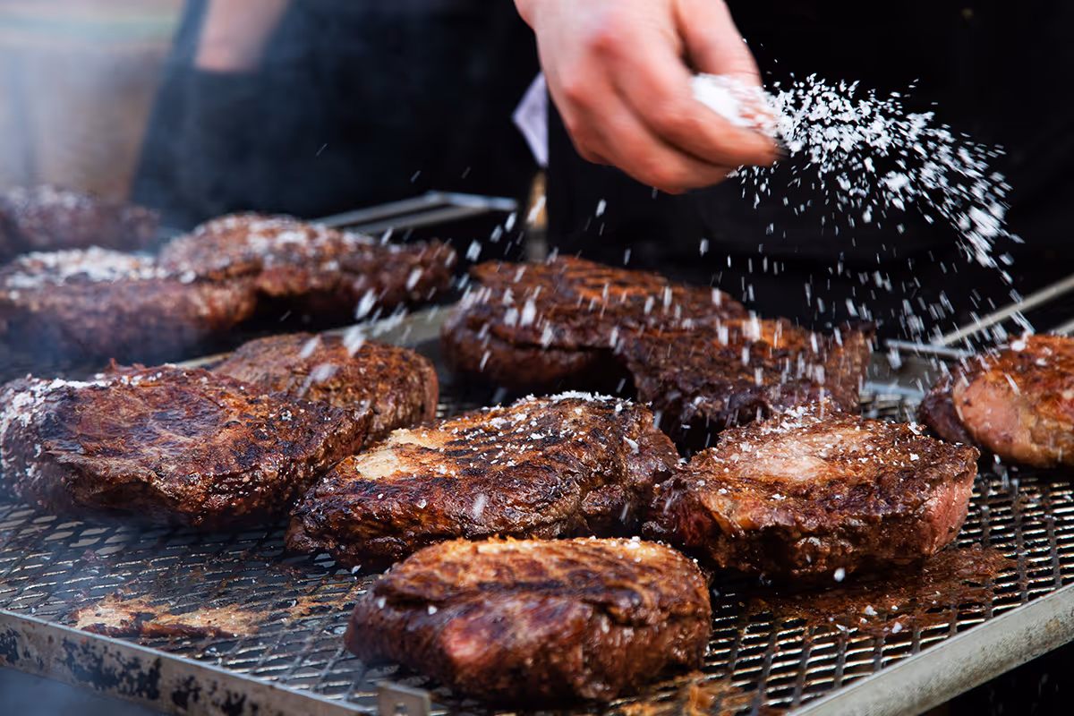 Hand sprinkling salt on grilled steaks cooking on an outdoor barbecue grill.