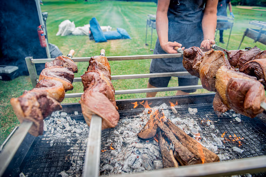 Close-up of meat roasting on skewers over an open fire pit in a grassy outdoor setting.