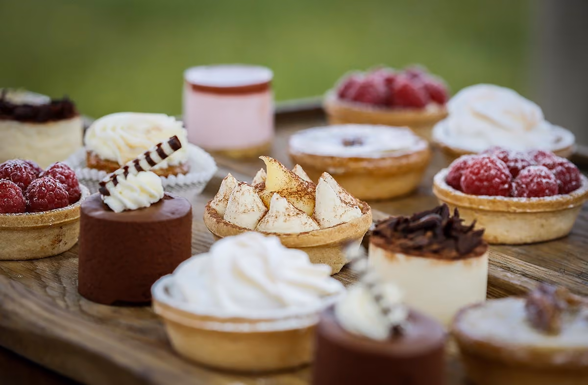 Wooden platter with assorted small dessert tarts including raspberries, cream, and chocolate toppings.