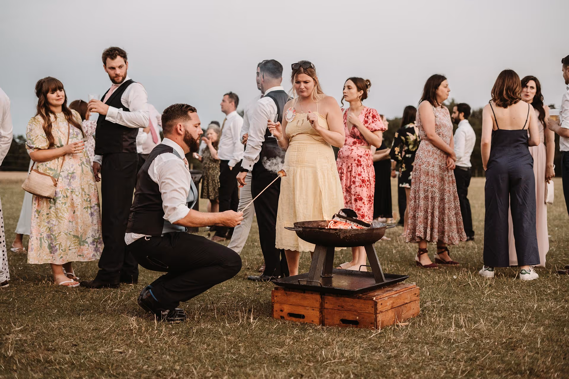 A group of people outdoors at a social event around a fire pit, one man roasting food on a stick.
