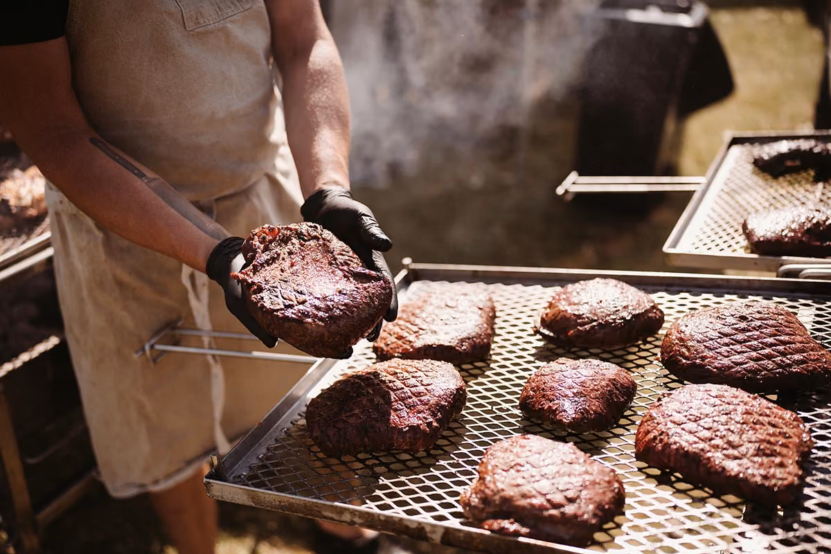 Person in apron holding a smoked brisket slab above a grill with several smoked meat pieces.