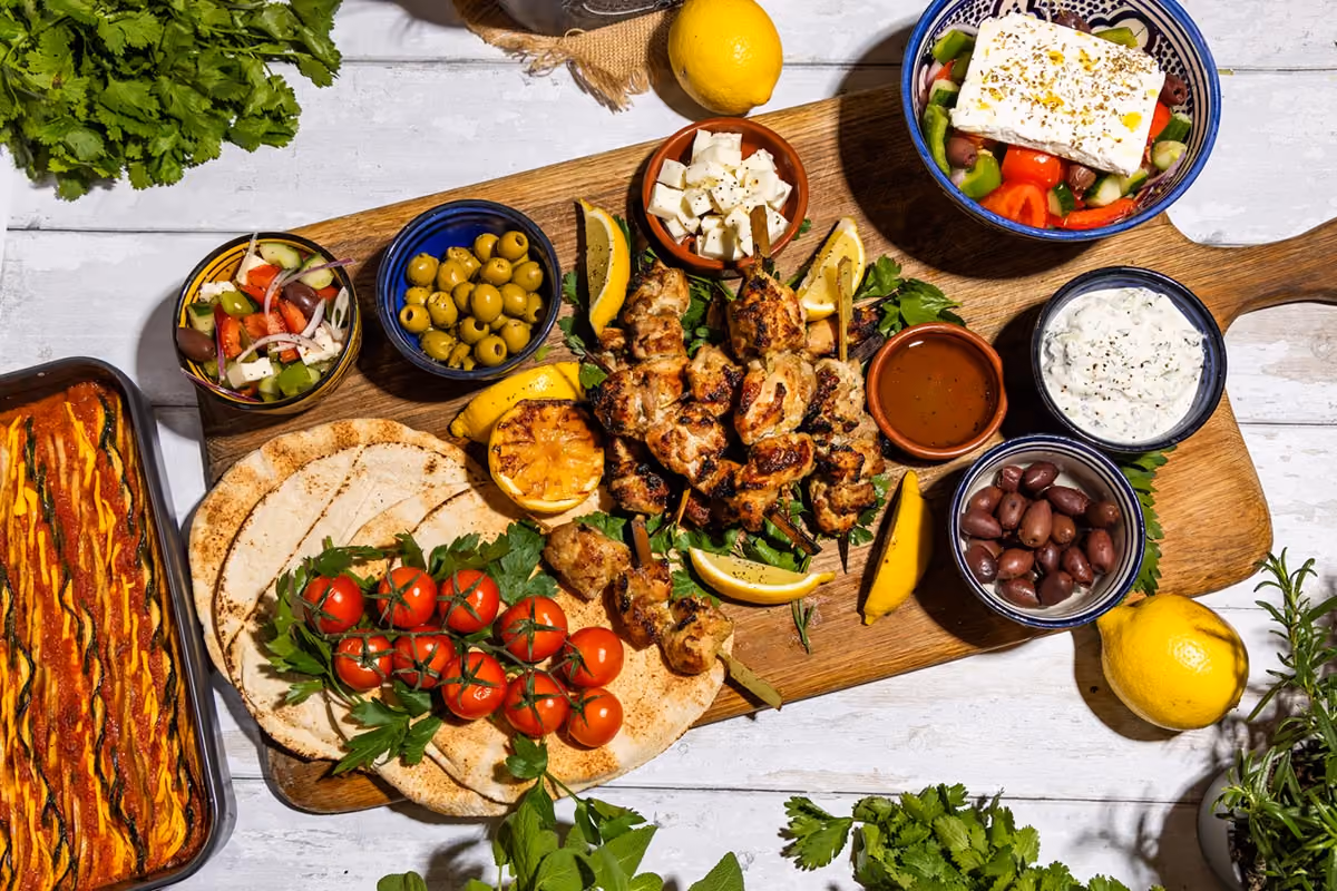 Wooden board with grilled chicken skewers, pita bread, cherry tomatoes, olives, feta, and dips.
